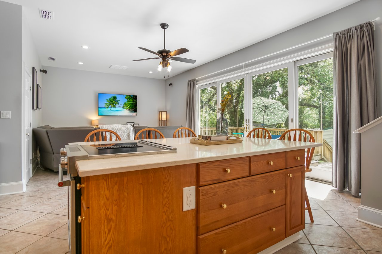 Spacious kitchen with a wooden island countertop, stools, and a ceiling fan. Large windows offer a view of greenery, creating a serene atmosphere.