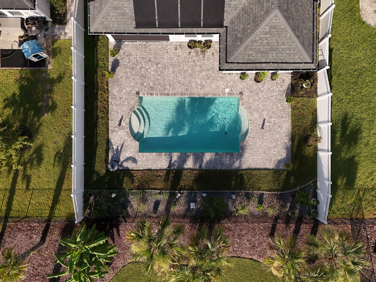 Aerial view of a backyard pool with a stone patio, white fencing, and landscaped lawn.