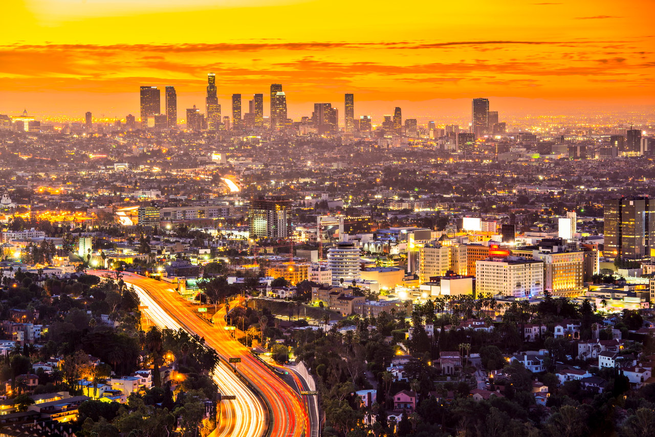 Aerial view of the Los Angeles skyline at sunset with downtown skyscrapers, city lights, and surrounding neighborhoods.