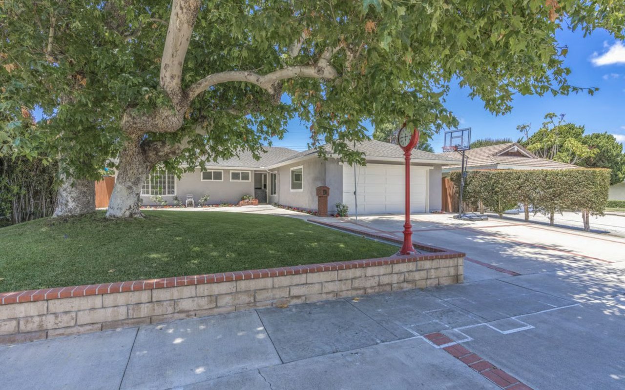 A grey suburban house with a large green lawn, a brick retaining wall, and a driveway with a basketball hoop.