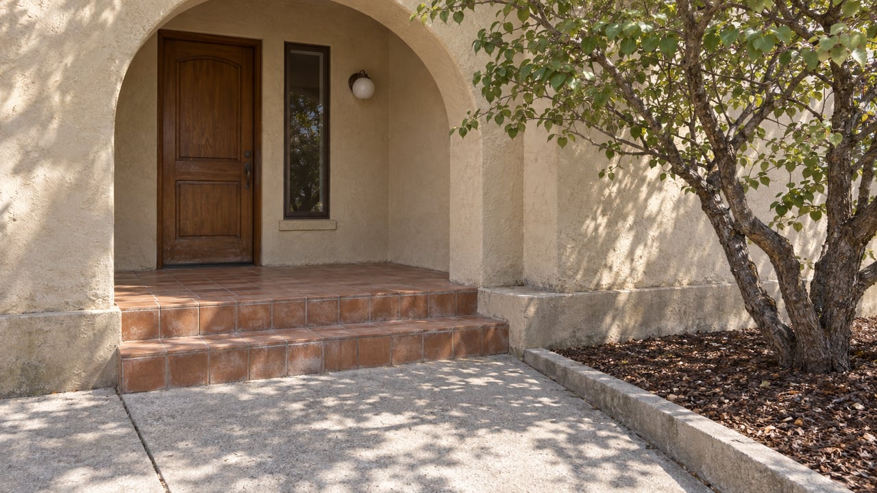 Sunlit stucco entry with a recessed wood door, worn terracotta steps, and a mature western redbud beside the concrete approach.