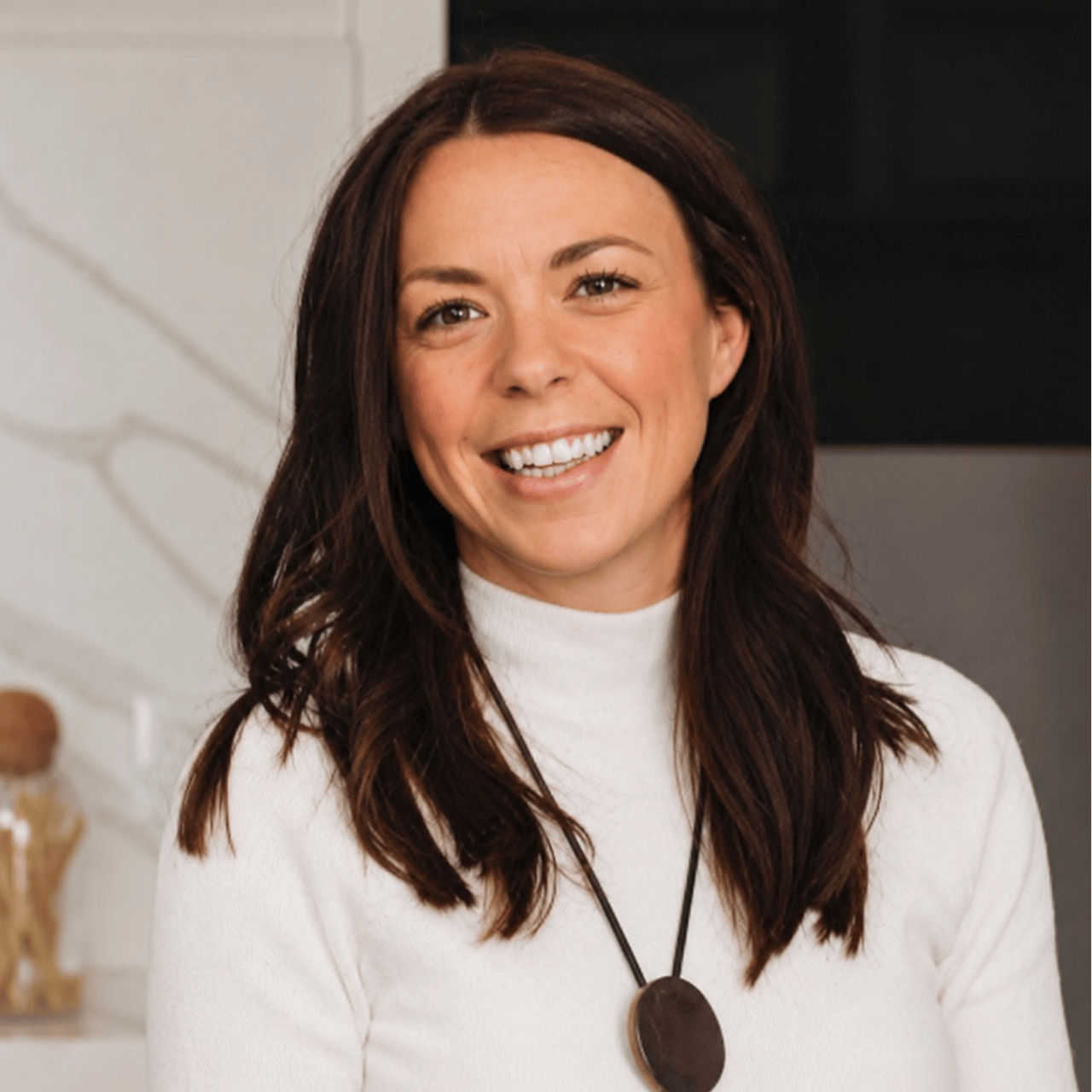 Smiling woman with long dark hair wearing a white sweater and round pendant necklace