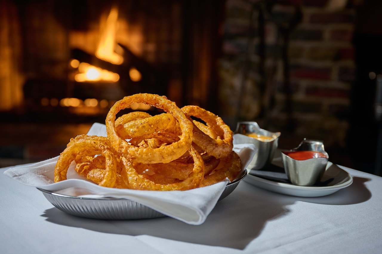Golden onion rings in a basket with dipping sauces beside a glowing fireplace.