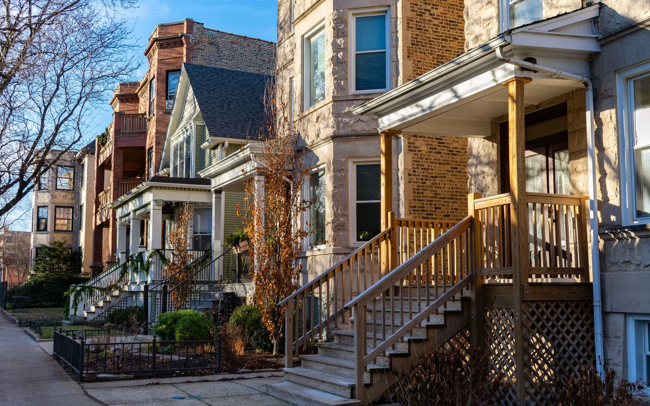 Residential street with brick and stone homes, wooden porches, and fenced front gardens.