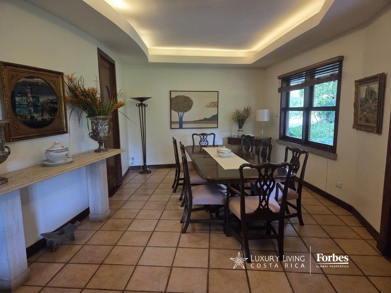 Dining room with a long wooden table, carved chairs, and a recessed ceiling with warm lighting.