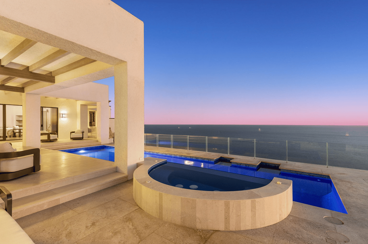 Oceanfront home terrace with a curved spa, swimming pool, and glass railing overlooking the sea at dusk.
