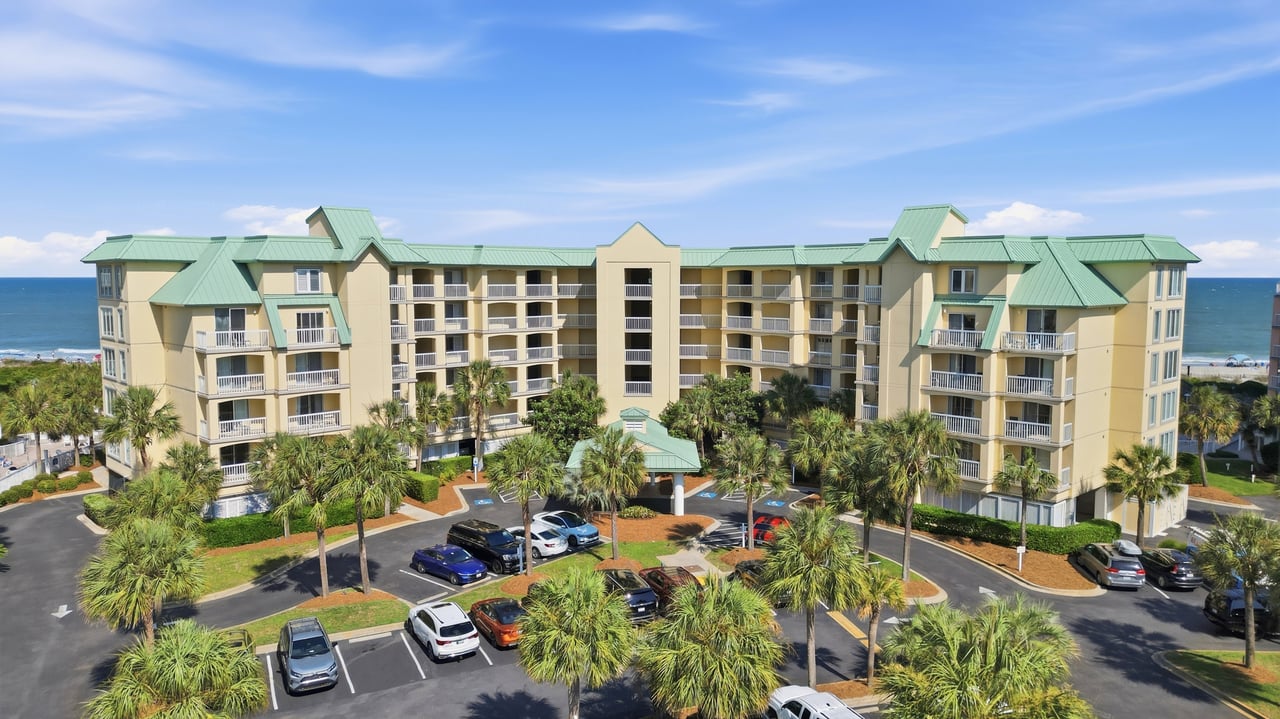 Coastal condominium complex with green roofs, palm trees, and ocean behind it.