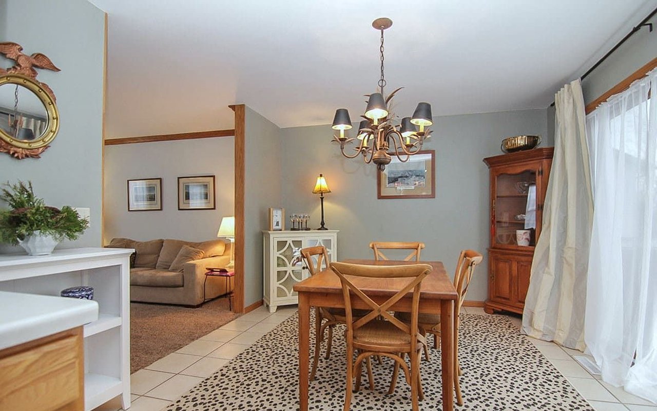 Furnished dining room with a wooden table, patterned rug, chandelier, and adjoining living room.