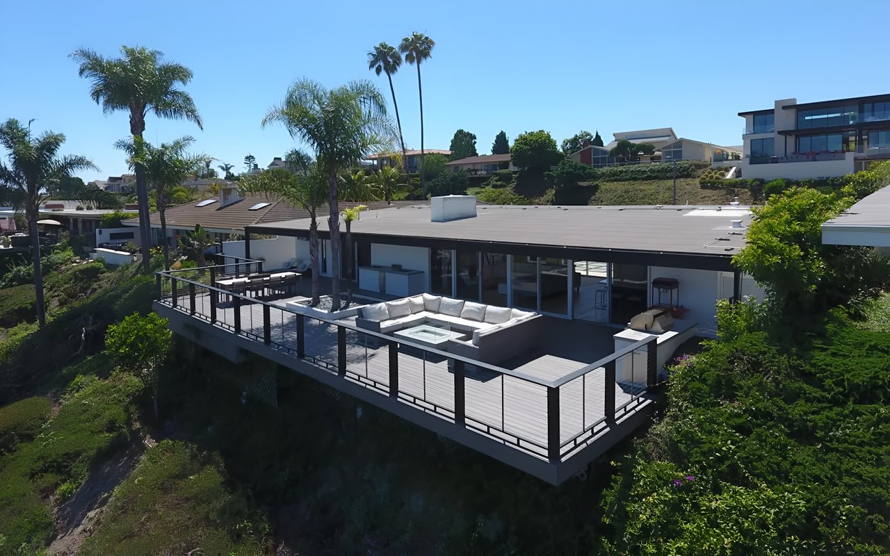 An aerial view of a modern house on a hill with a large deck, an outdoor seating area, and palm trees.