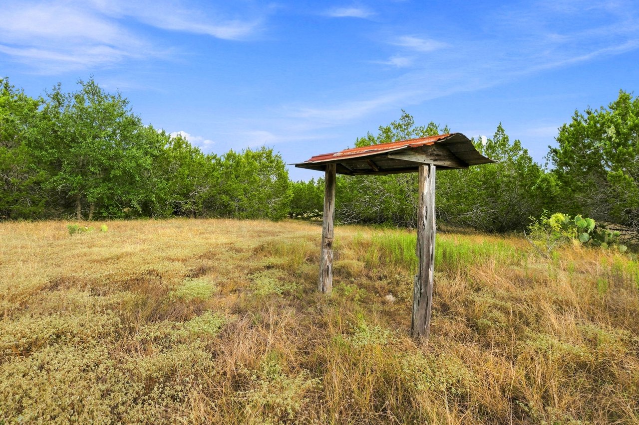 Texas Hill Country meadow with a rustic wooden shelter, dry grass, and dense trees.