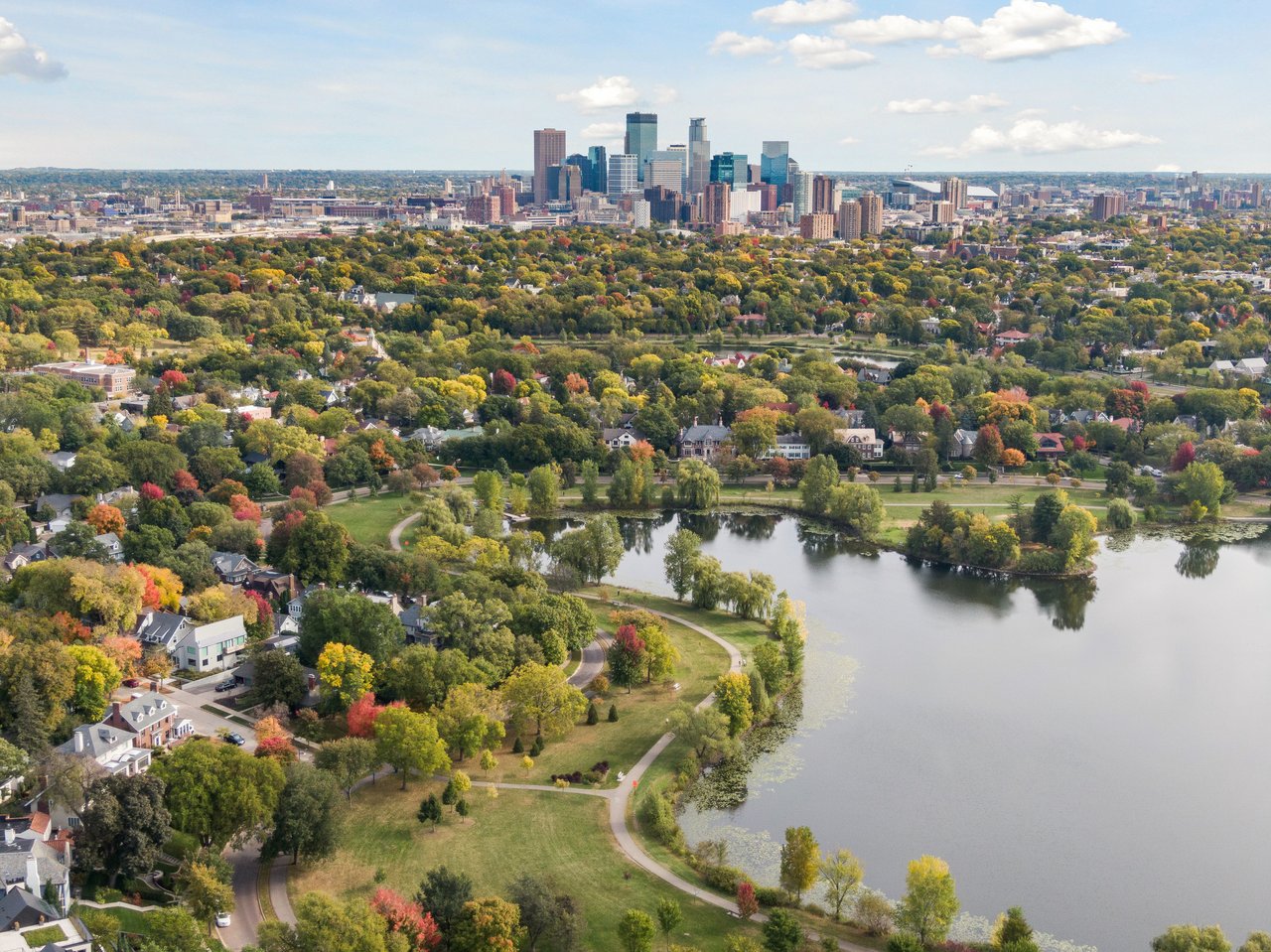 An aerial view of Lake of the Isles surrounded by lush green parks and residential neighborhoods, with the downtown Minneapolis skyline visible in the background, located in the vibrant Minneapolis lakes area, represented by the Cari Ann Carter Group.