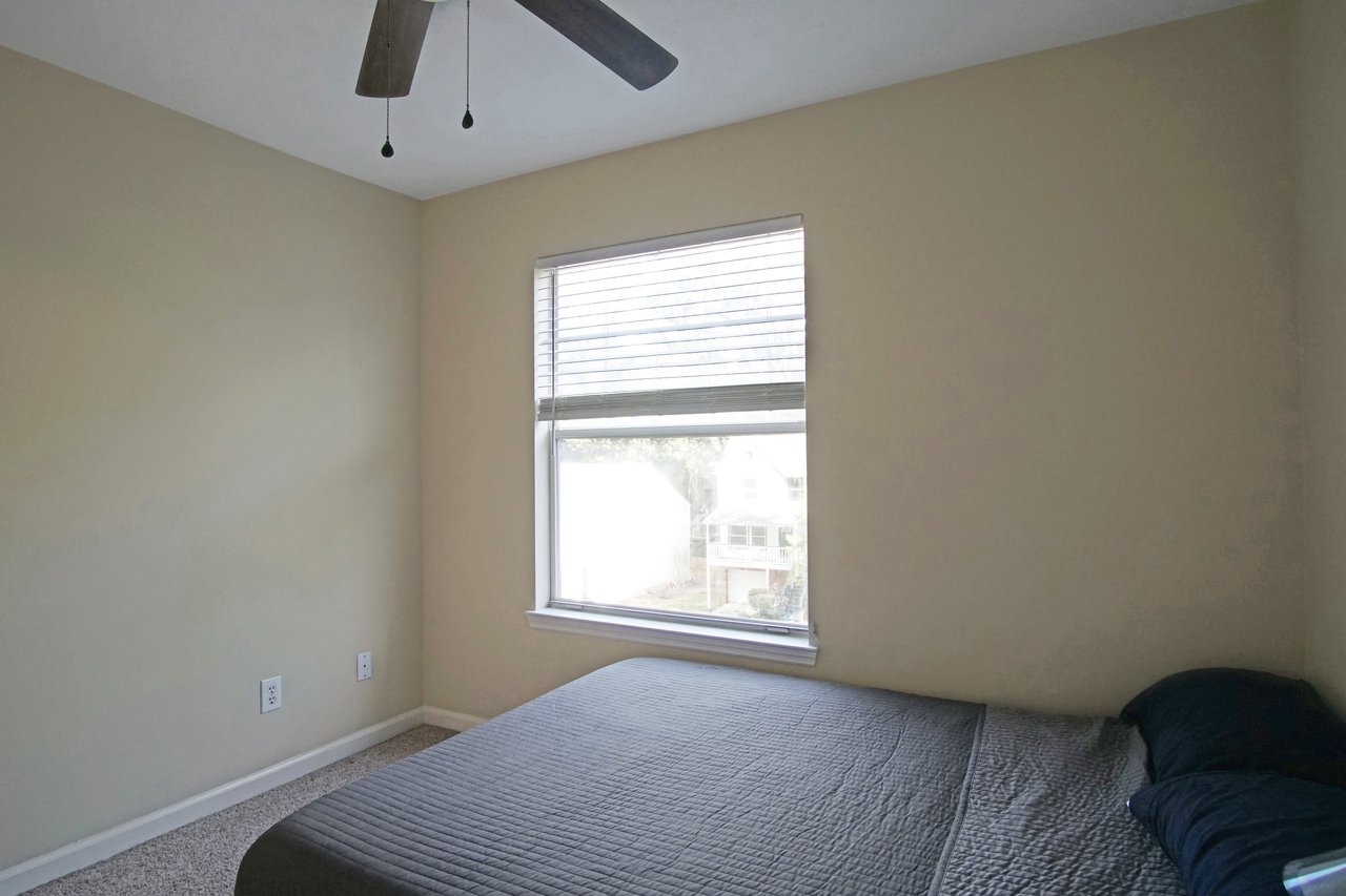 Bedroom at 529 Collinsford Road, Tallahassee, Florida, featuring beige walls, carpet flooring, and a ceiling fan with a light fixture. The room includes a bed with gray bedding positioned beneath a large window with blinds that let in natural light.