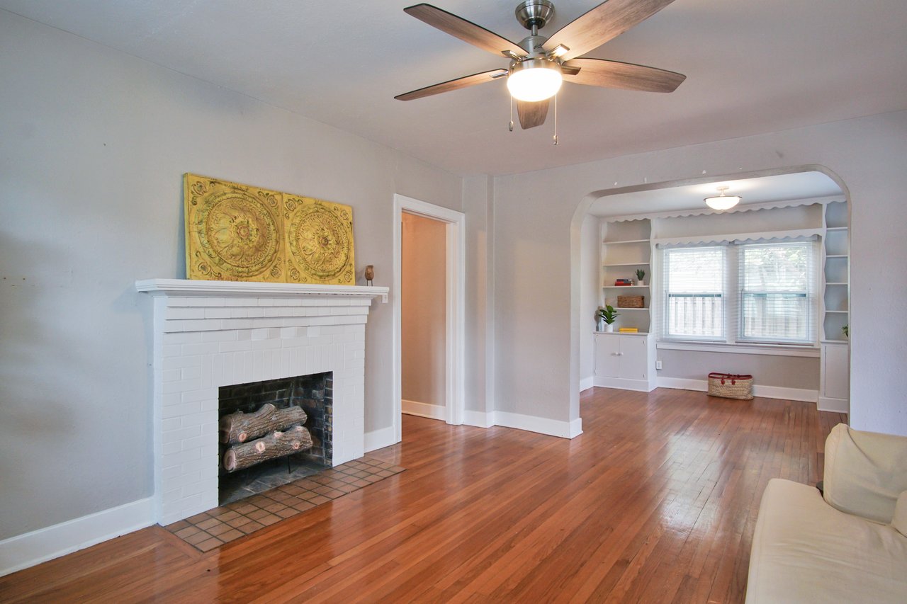 A cozy living room with wooden floors, a white brick fireplace adorned with yellow artwork, and a ceiling fan. A bright adjoining area features large windows.