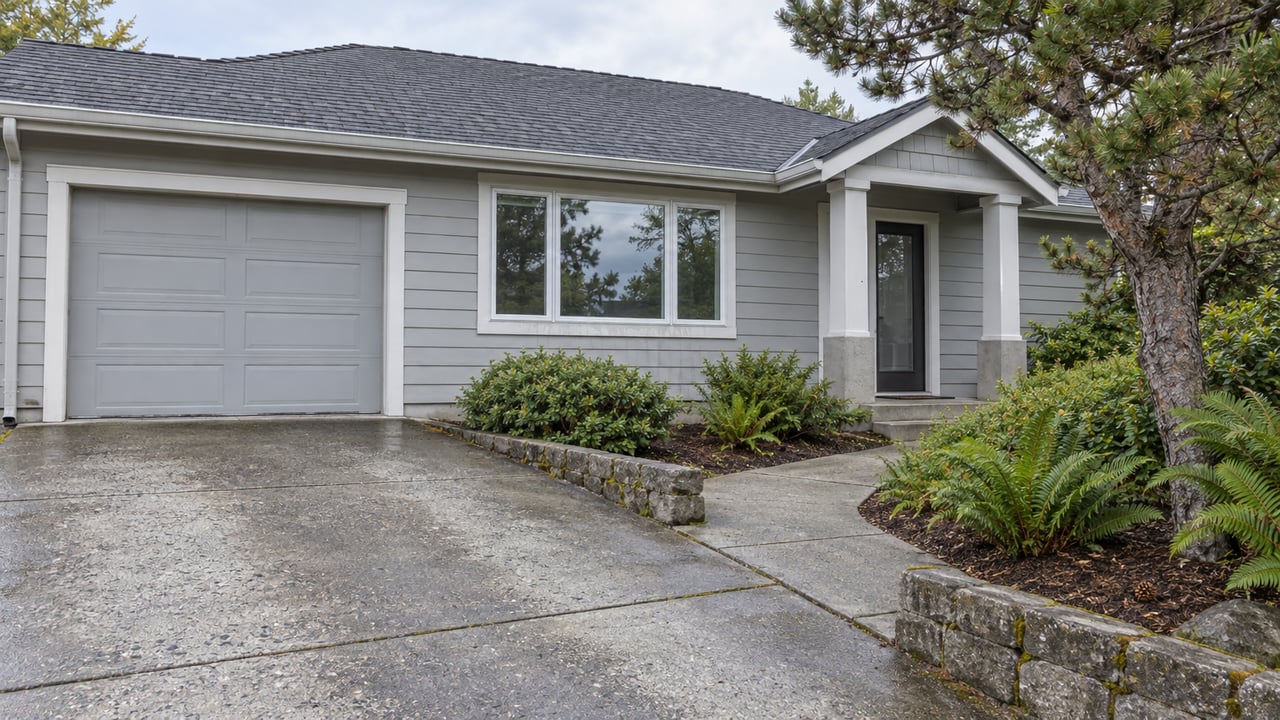 Light-gray gabled home with a concrete driveway, sheltered entry and wind-shaped pine beside a stone-edged planting bed.