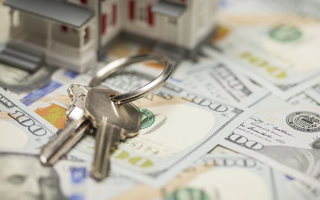 House keys on US $100 bills, with miniature house models in the background.