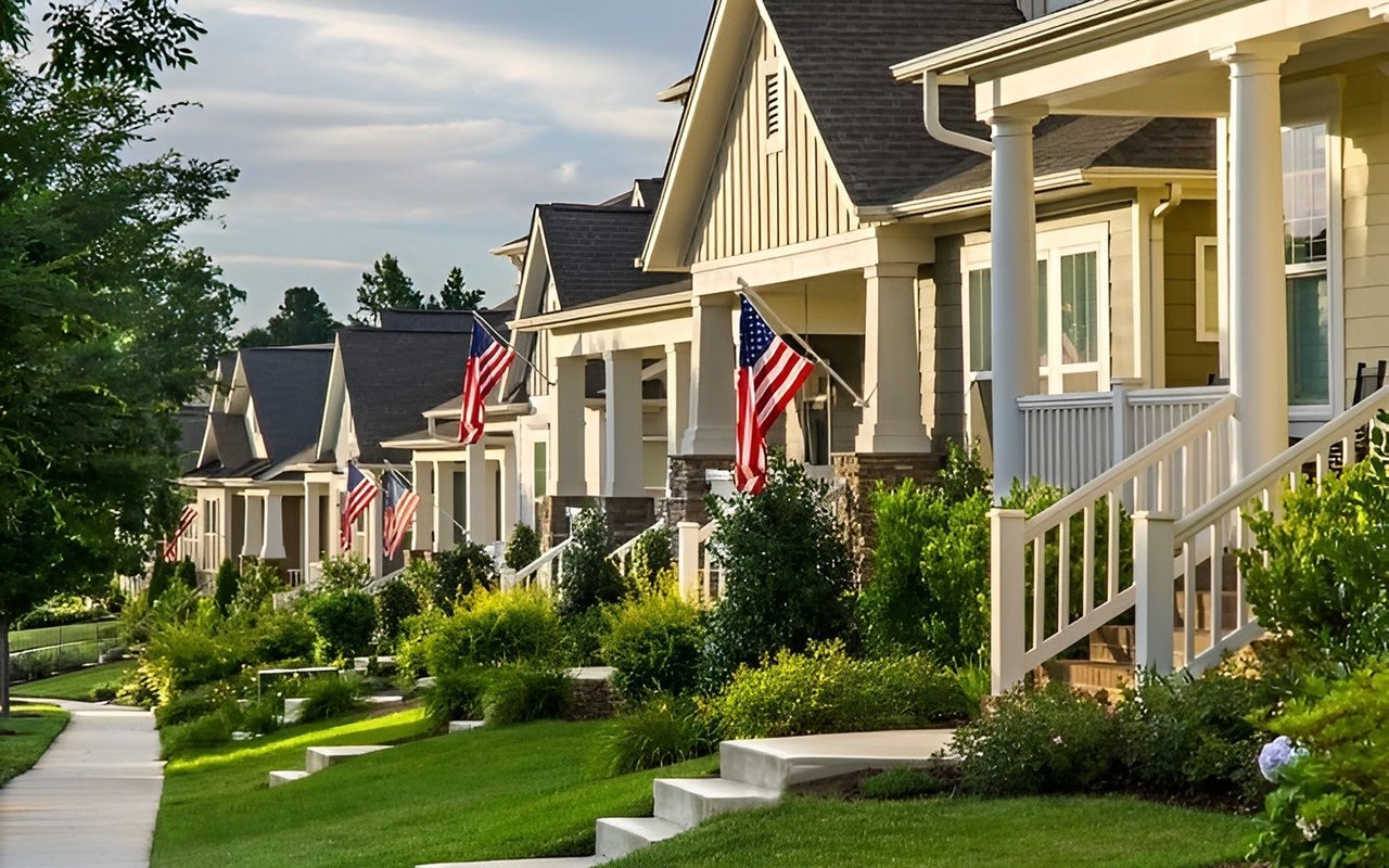 Suburban homes with covered porches and American flags along a landscaped sidewalk.