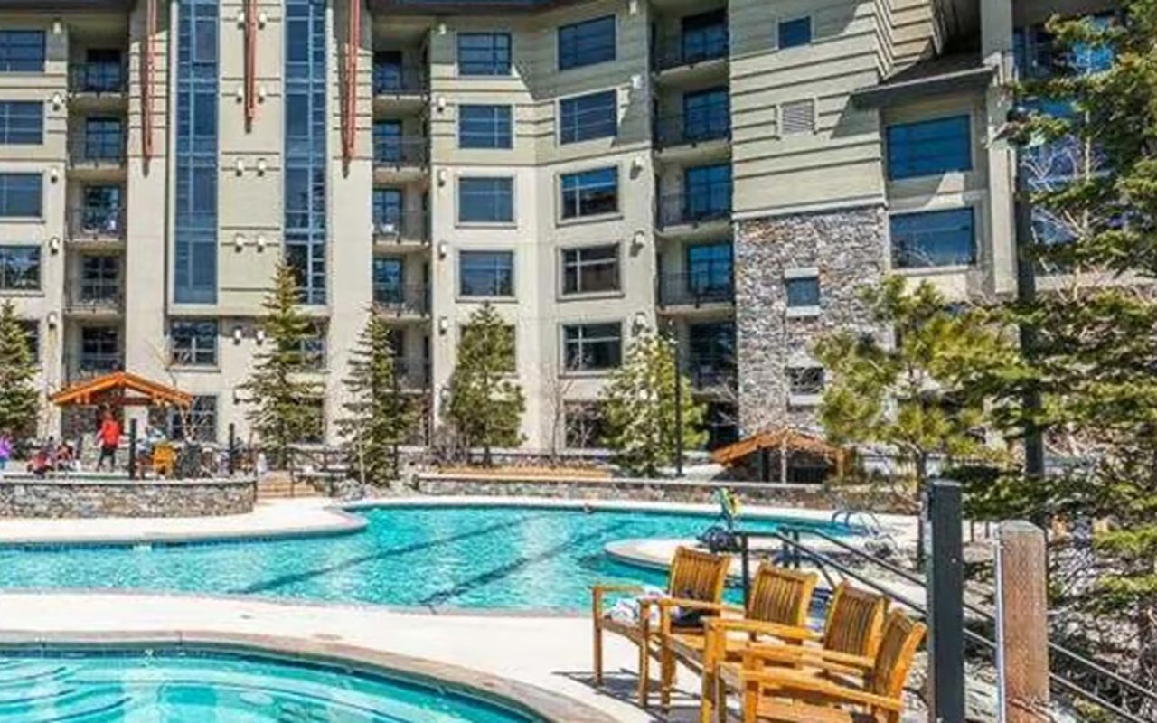 Condominium building overlooking a courtyard pool and hot tub, with wooden chairs beside the water.