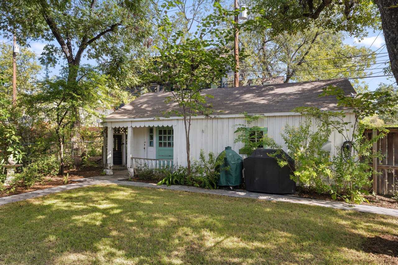 White cottage exterior with a teal door, shaded lawn, and mature trees.