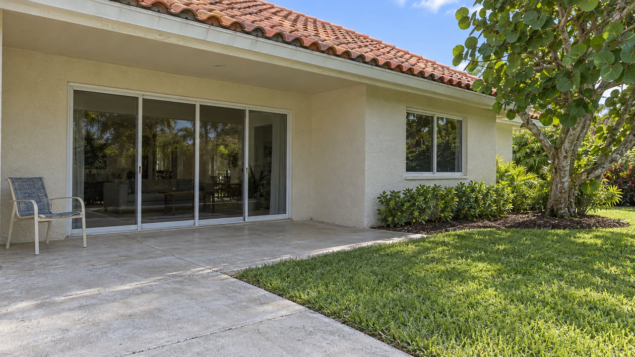 Sunlit concrete patio and lawn beside a cream stucco home with clay roof tiles, closed sliding glass doors and a sea-grape tree.