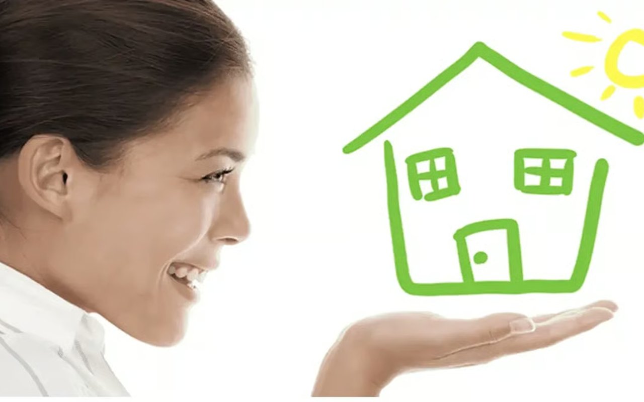 Smiling woman presents a green house drawing on her palm beneath a yellow sun.