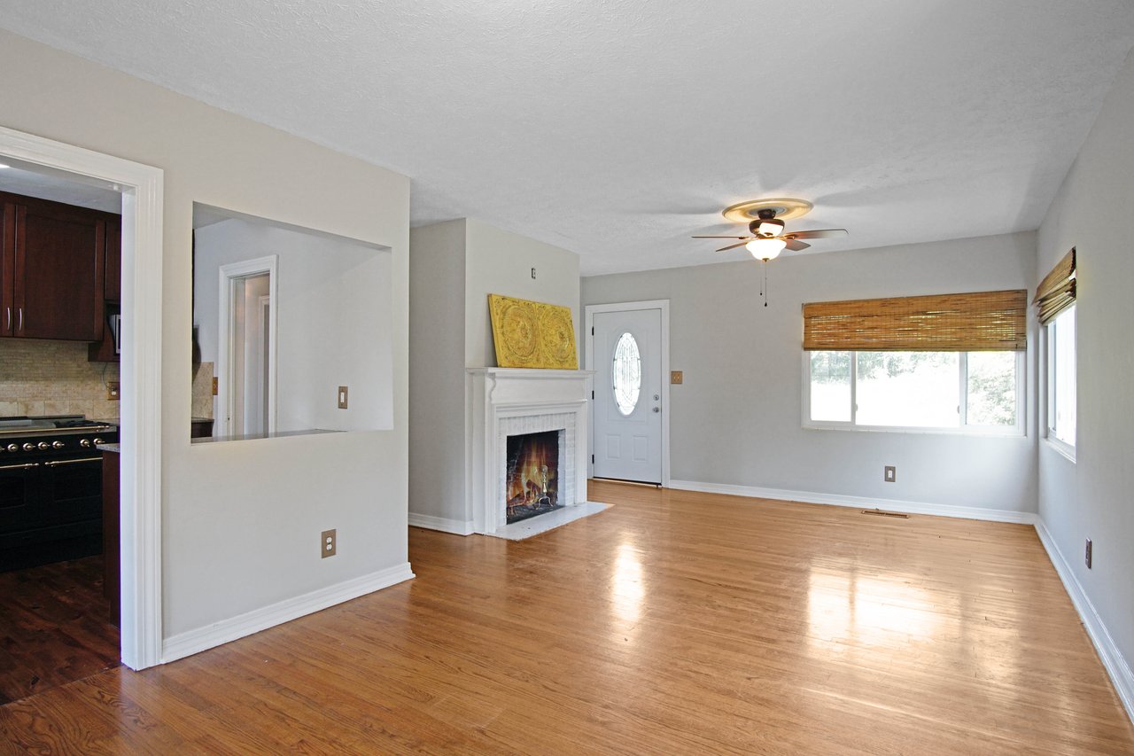 Bright living room with wooden floors, beige walls, and a ceiling fan. Features a white fireplace, French doors, and a window with bamboo shades. Cozy and inviting.