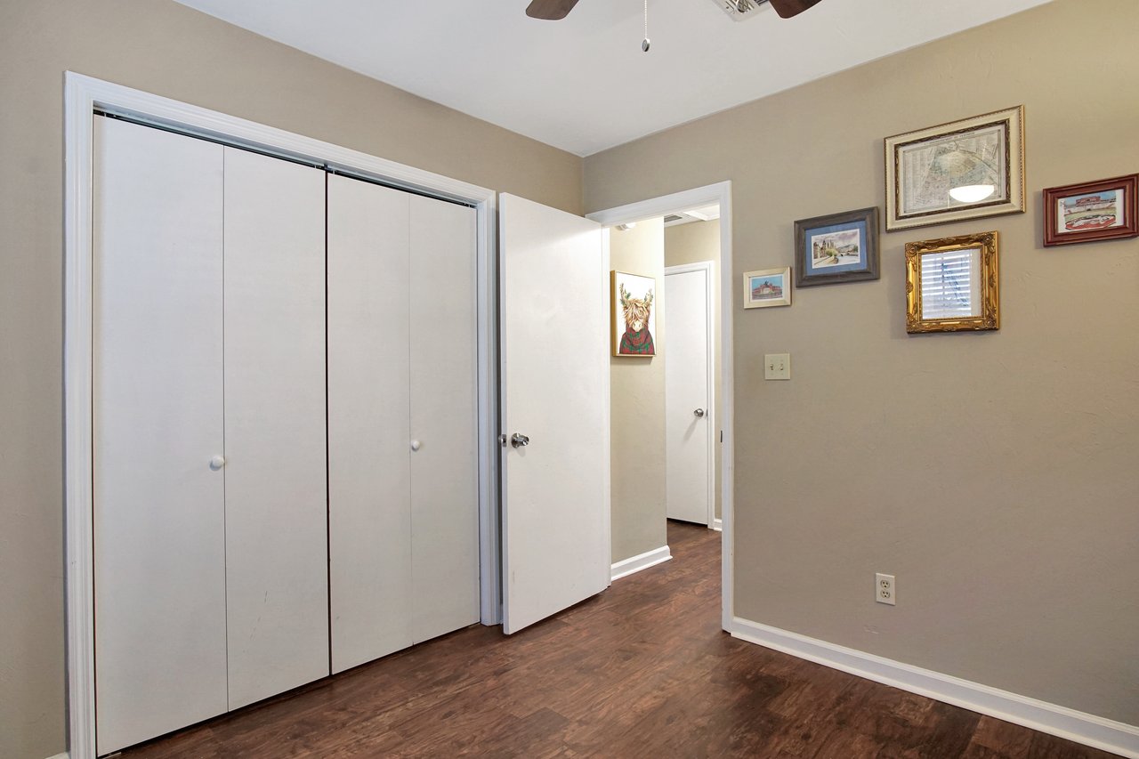 A cozy room with beige walls and wooden floors. A door is ajar, revealing a hallway. There are several framed artworks on the wall, adding warmth.