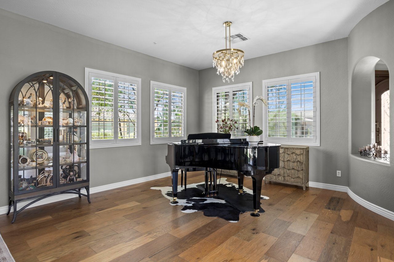 A formal dining room with gray walls, wood floors, plantation shutters, a chandelier, a black grand piano, and a display cabinet.