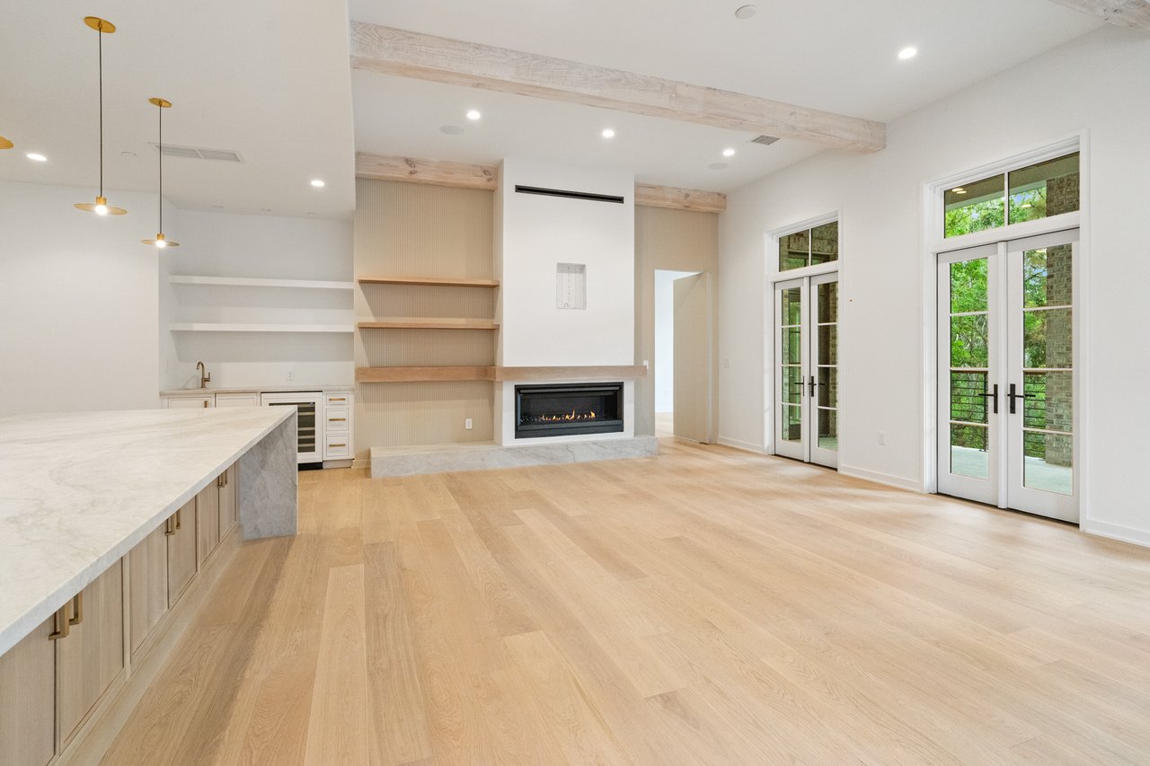 Open-plan living room with pale wood floors, built-in fireplace, kitchen island, and tall French doors.