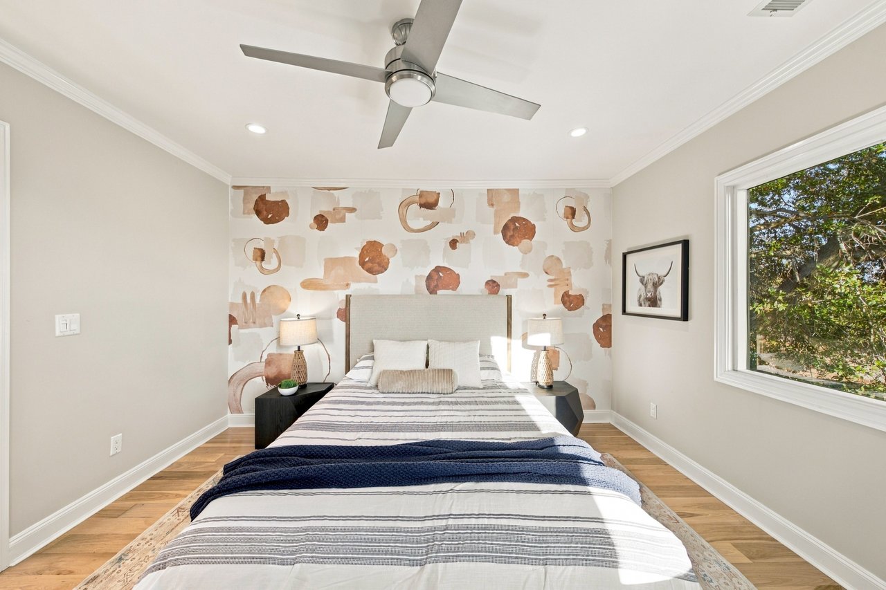 Bedroom with a patterned accent wall, centered bed, and large window overlooking trees.