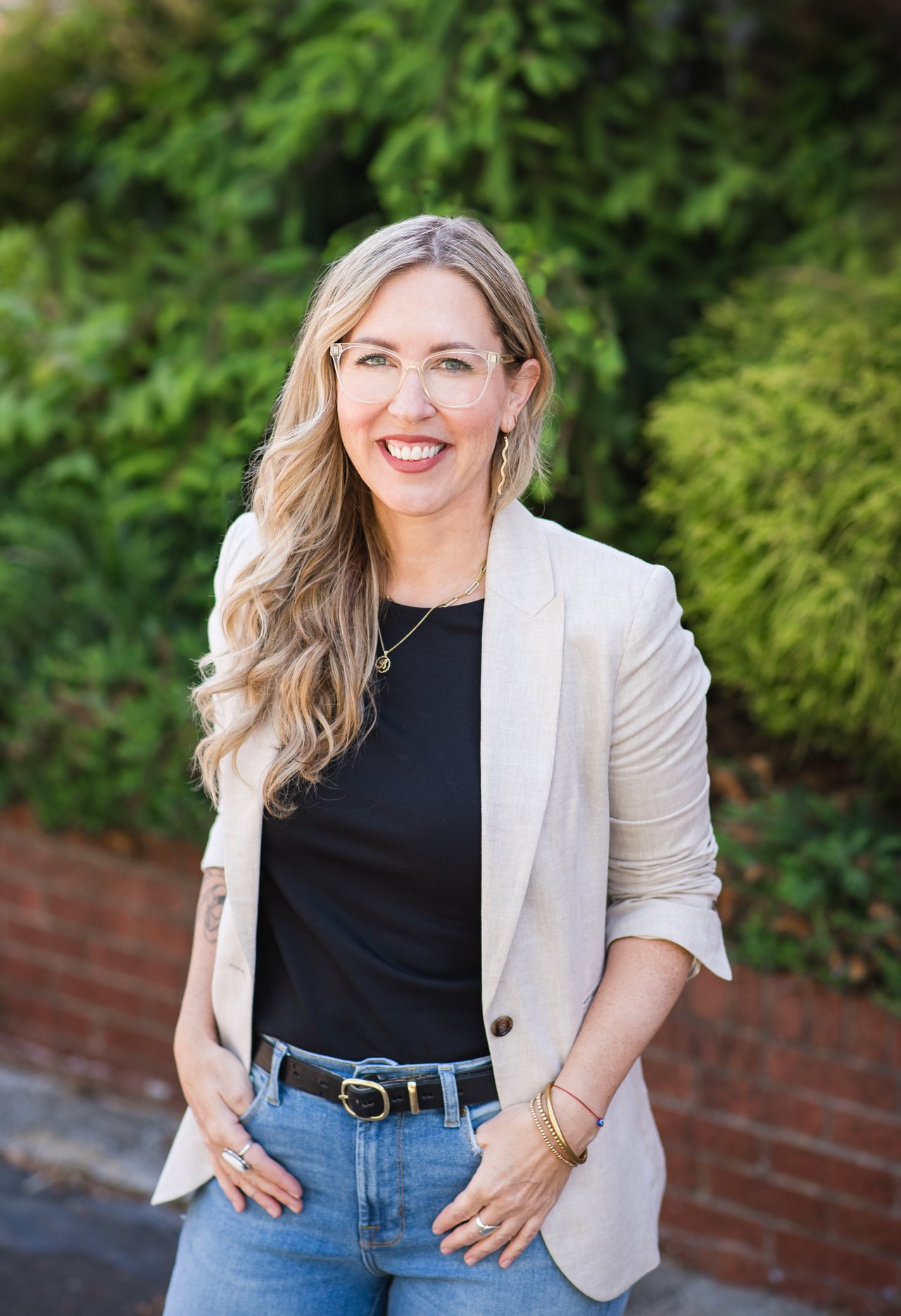 Annie Blue smiling outdoors in a light blazer, with leafy greenery and a brick wall behind her.