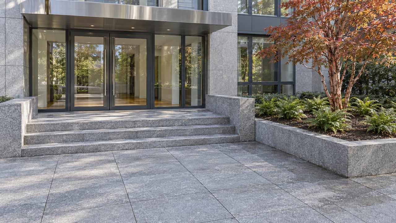 A contemporary glass condominium entrance beyond a broad granite forecourt, with a copper-leaved paperbark maple in a raised stone planting bed.