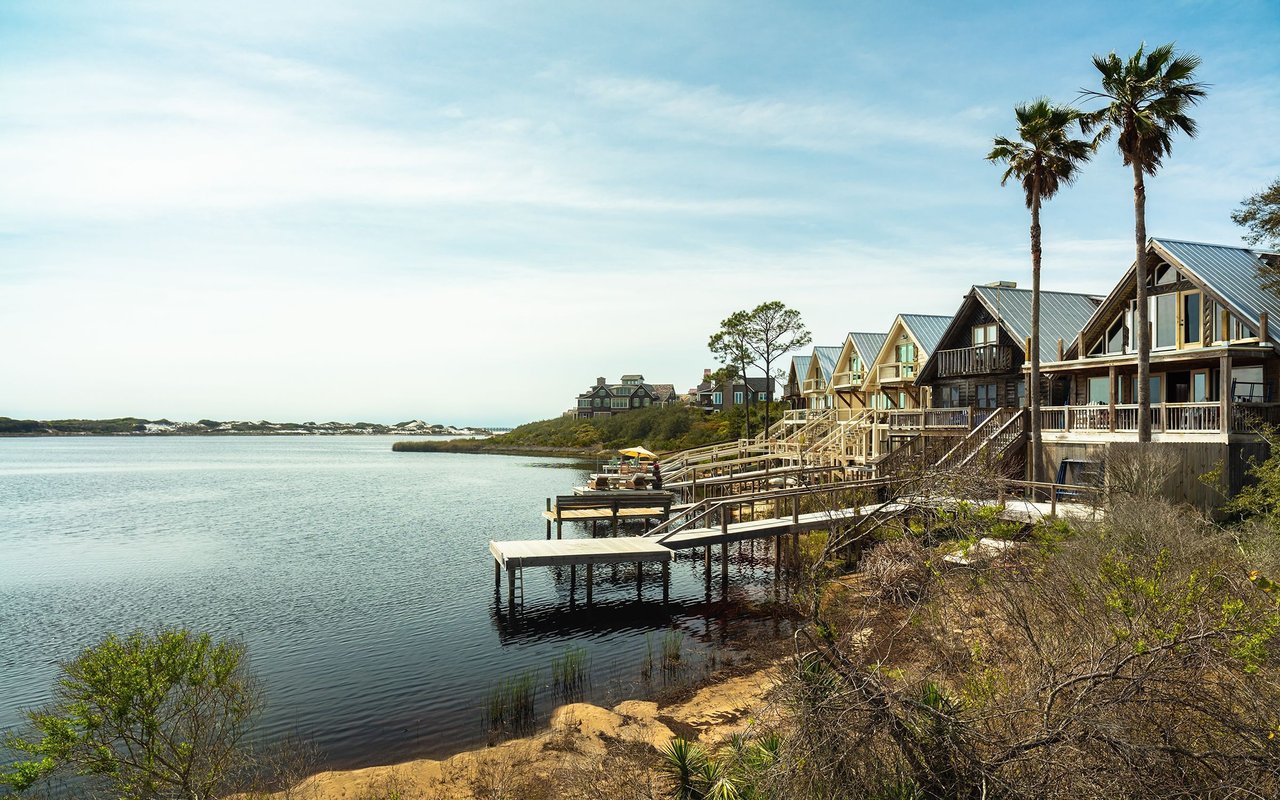 Waterfront homes with wooden decks and docks along a calm bay, framed by palm trees.