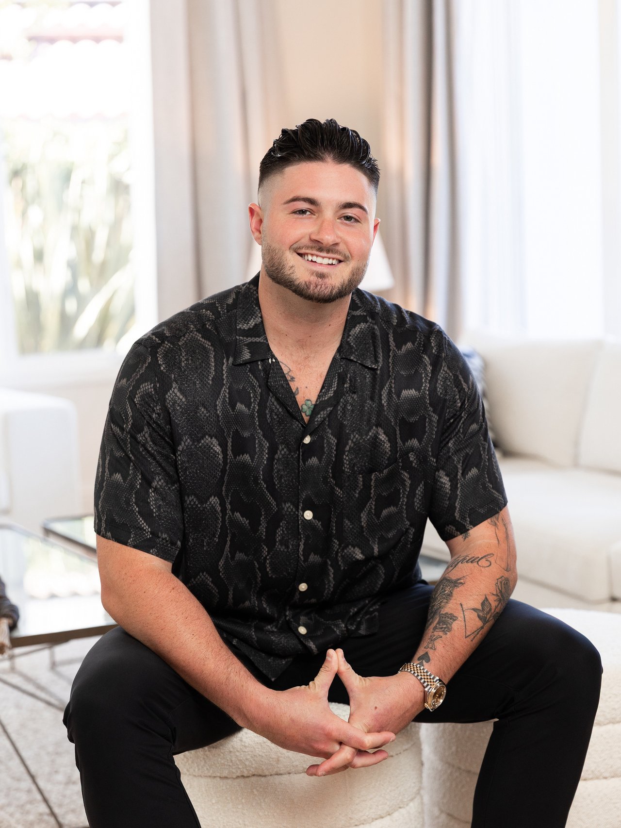 Smiling man in a black patterned shirt seated with clasped hands in a bright living room