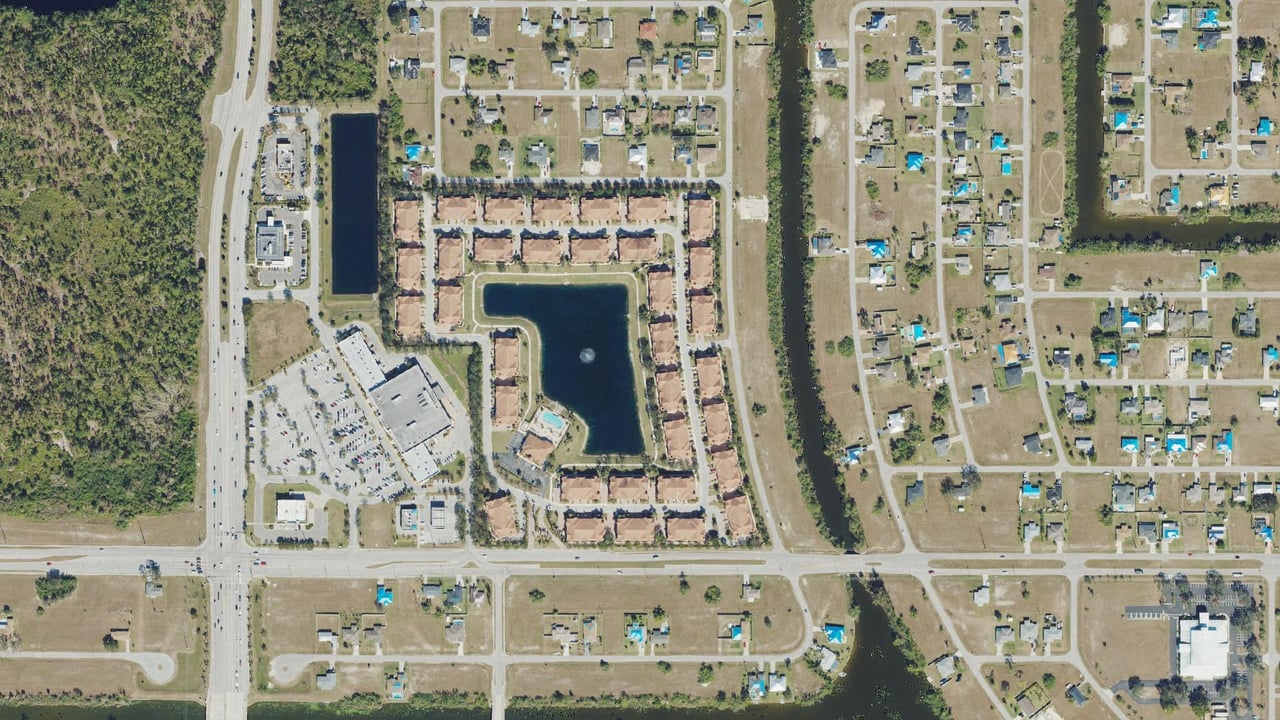 Aerial view of Concordia in Cape Coral, Florida: 34 two-story condominium buildings around a five-acre lake with a fountain and pool, beside the Publix center on Del Prado Boulevard
