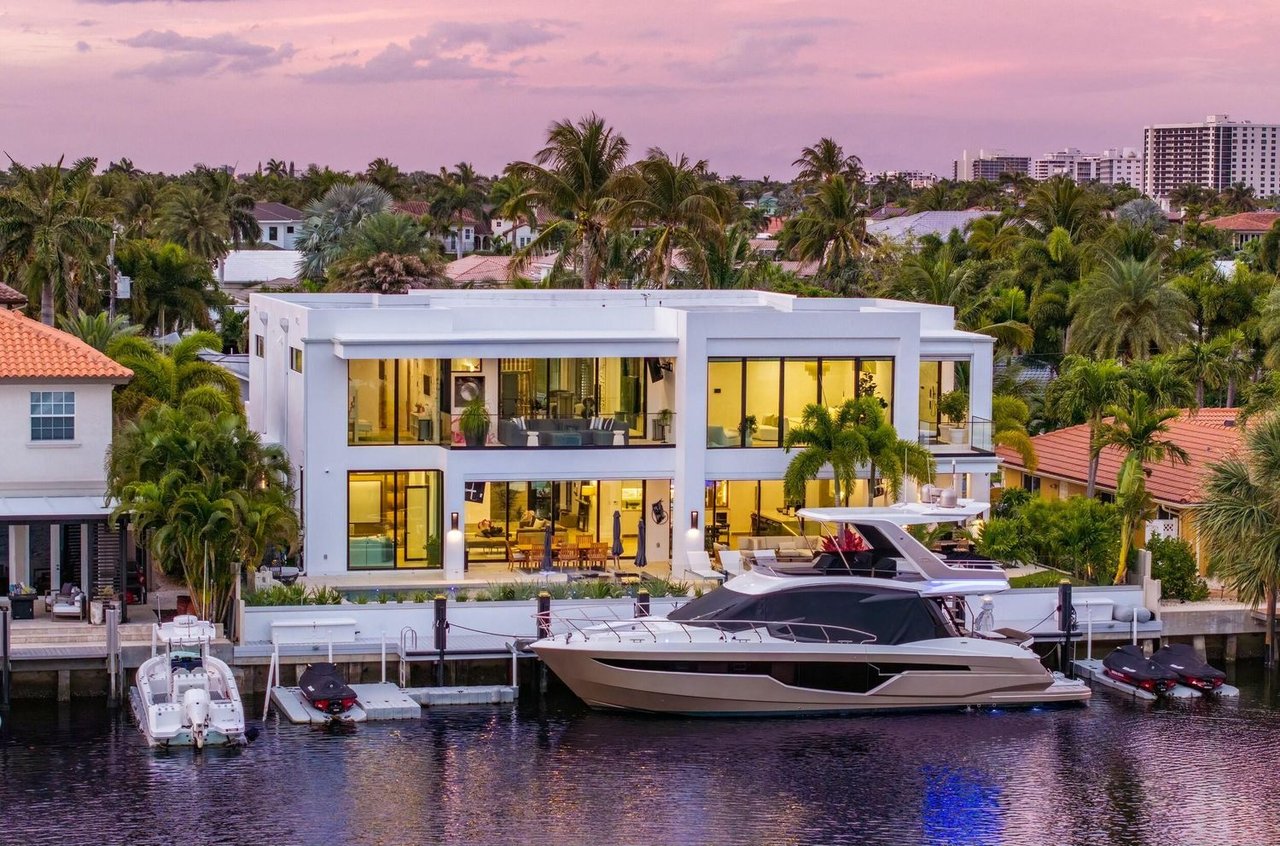 Modern waterfront home glowing at dusk, with palm trees, a canal, and a large yacht at its dock.