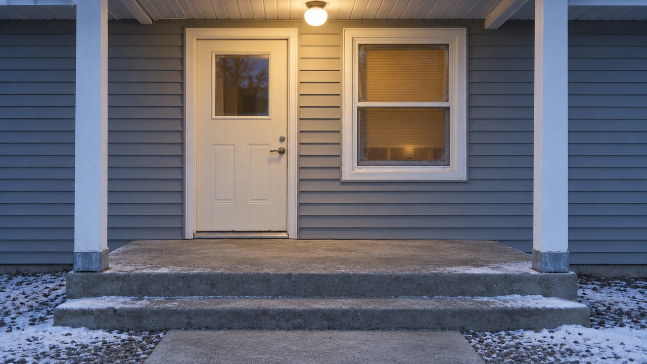 A modest gray-sided house entry at blue hour, with a warm porch light, closed insulated door and snow along the concrete steps.