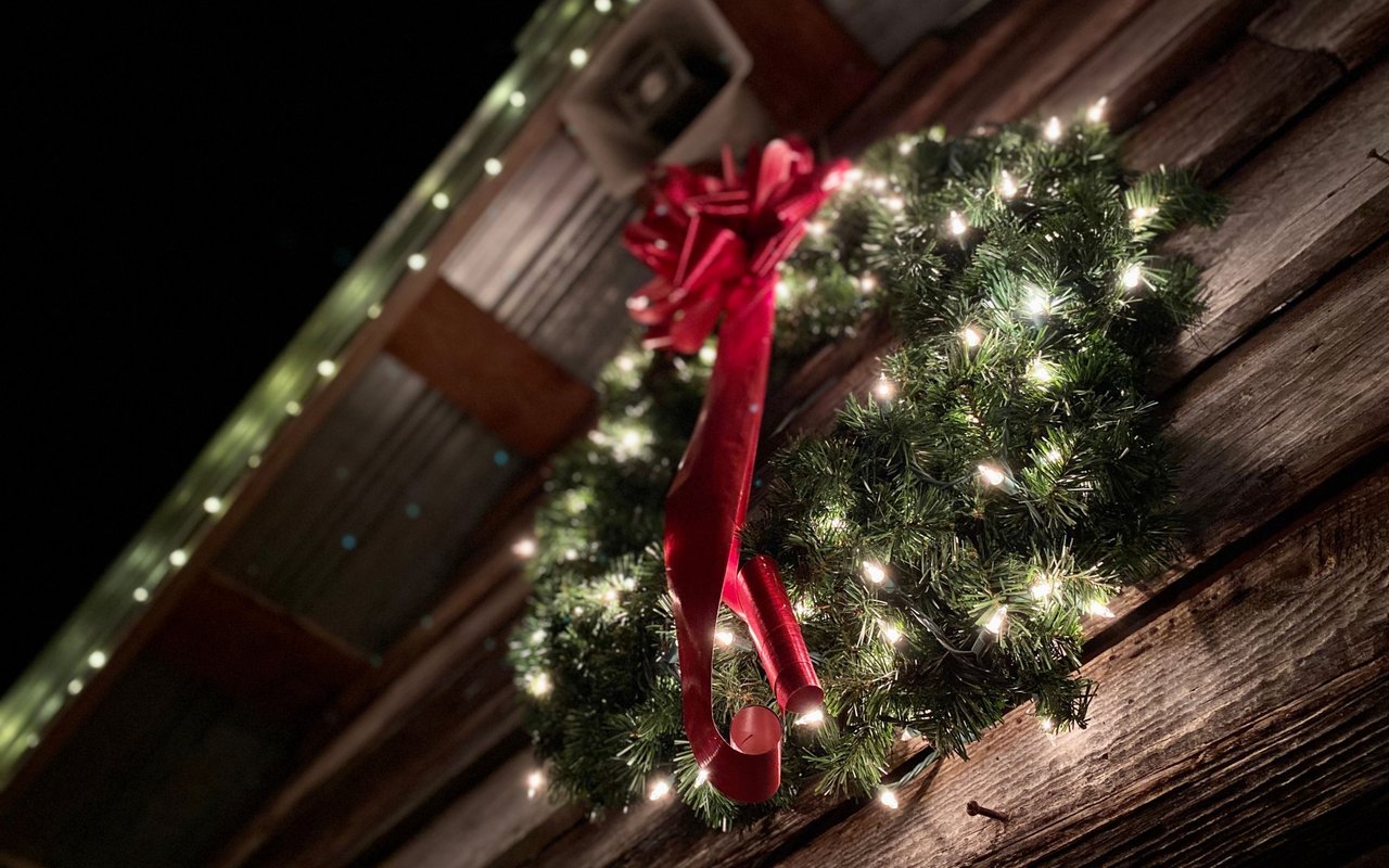 Lighted Christmas wreath with a large red bow on rustic wooden siding