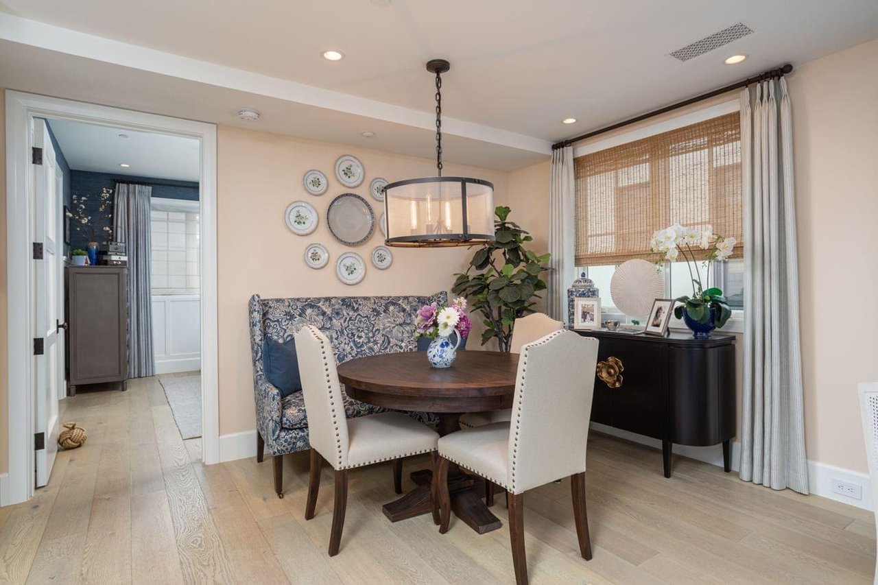  A bright dining room with a round wooden table, a banquette bench, and two chairs. A collection of plates hangs on the wall.