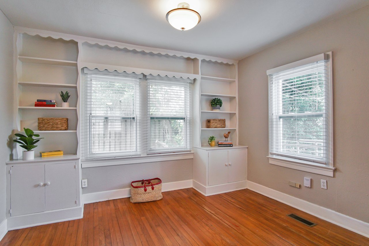Bright room with white walls and wooden floor, featuring built-in shelves around a window. Decor includes plants, wicker baskets, and books, creating a cozy, inviting atmosphere.