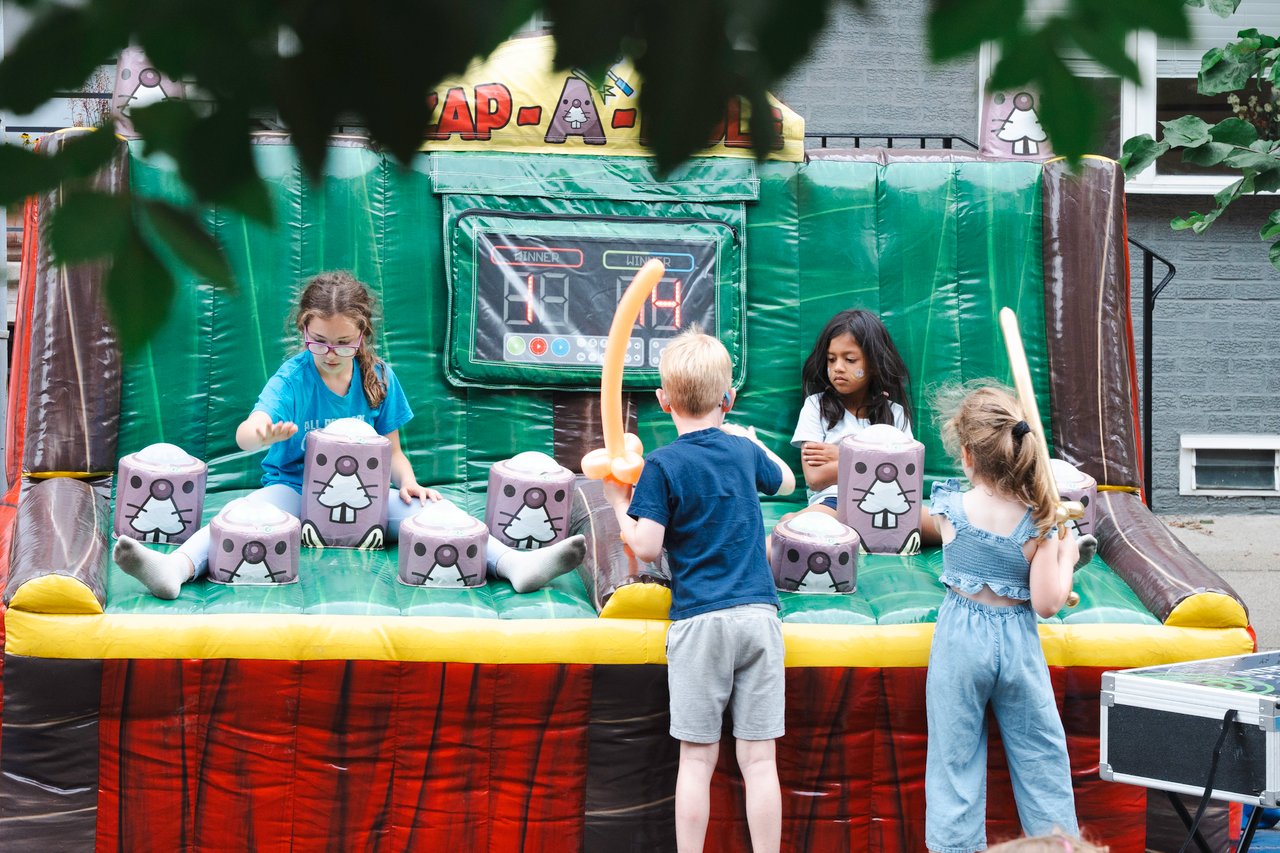 Children play at an inflatable game at a neighborhood block party, with one holding a balloon sword.