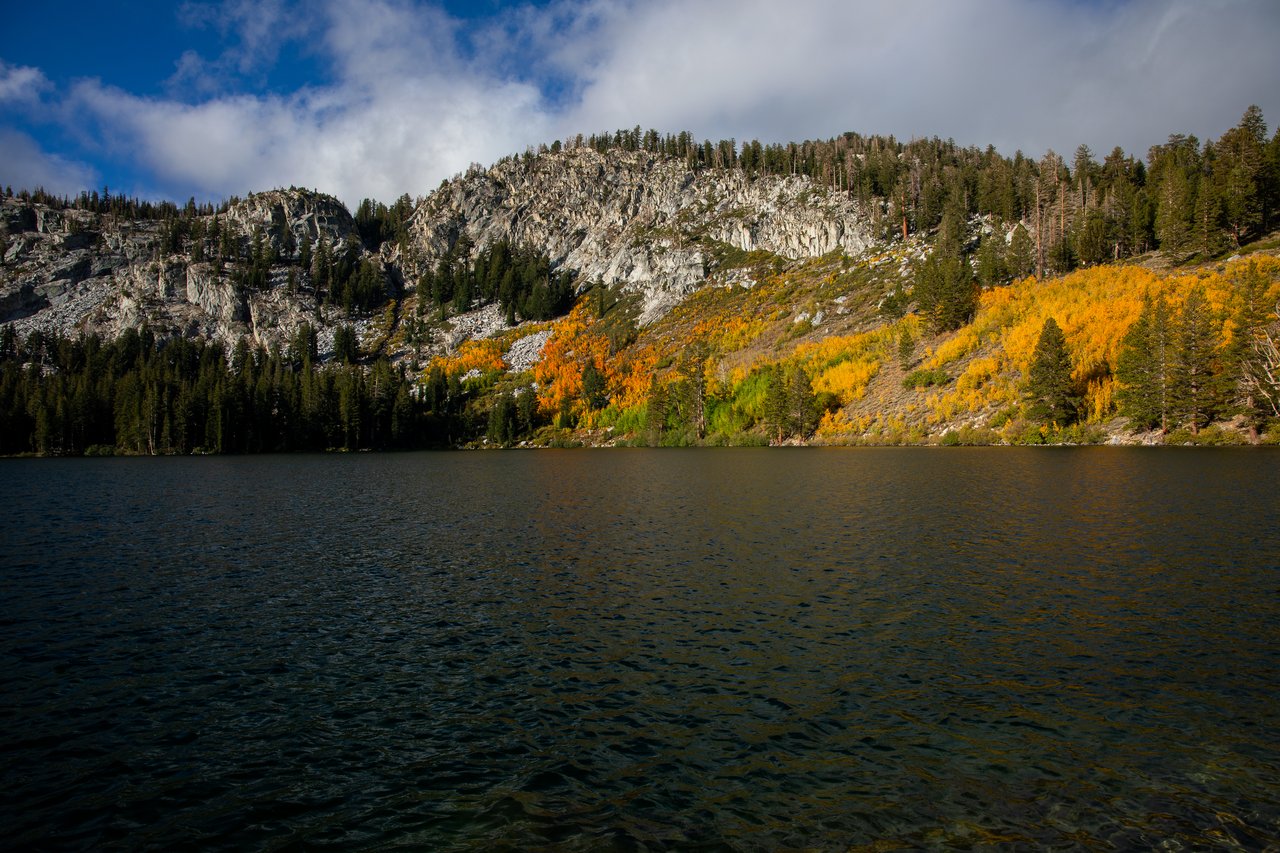 Alpine lake beneath rocky, pine-covered slopes streaked with golden autumn foliage.
