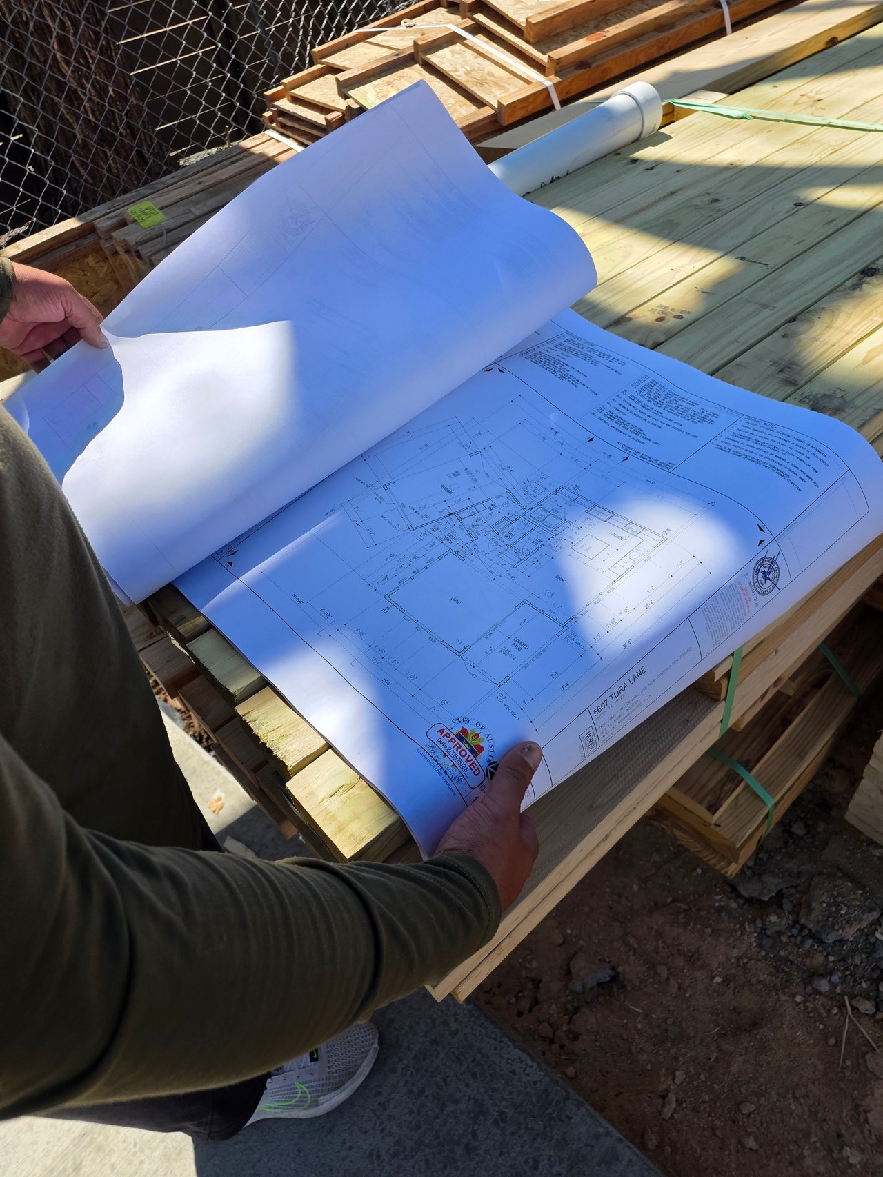 A person reviews City-approved building plans spread across lumber at a construction site.