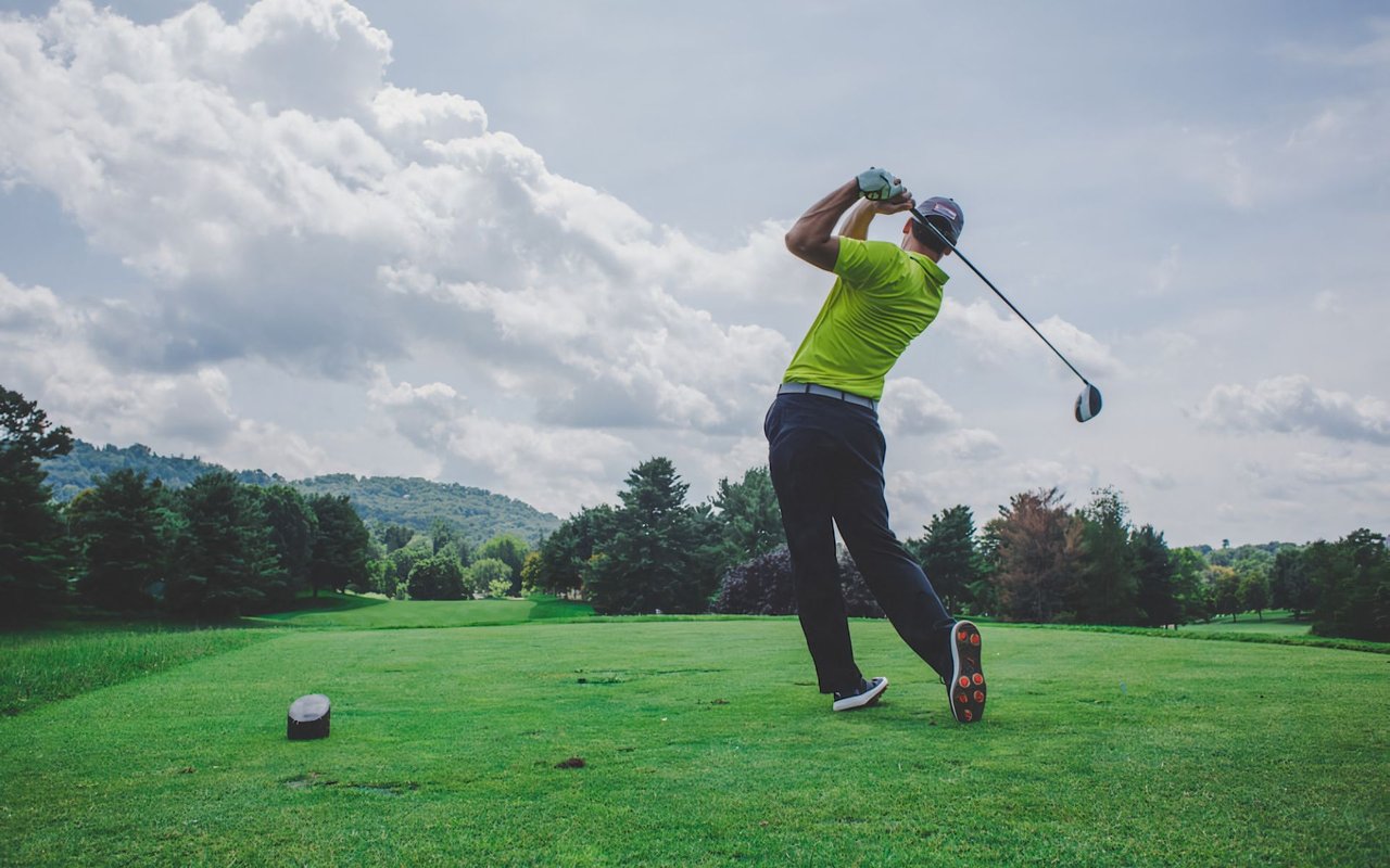  A male golfer in a bright yellow-green polo shirt, dark pants, and a cap is captured mid-swing, having just hit the golf ball.