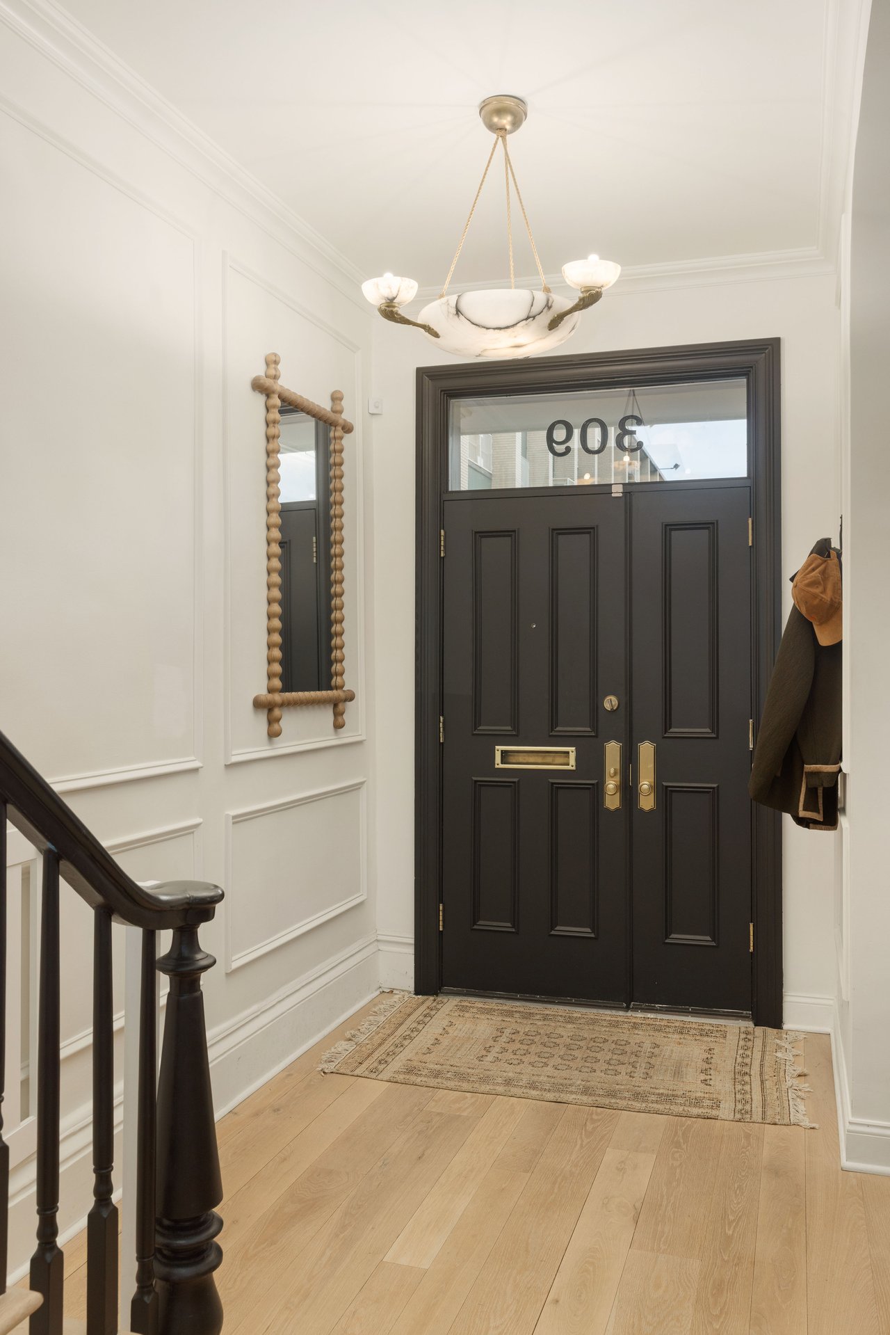 Elegant entryway featuring double black paneled doors with a transom window, white wainscoting, light oak flooring, and a decorative beaded mirror.