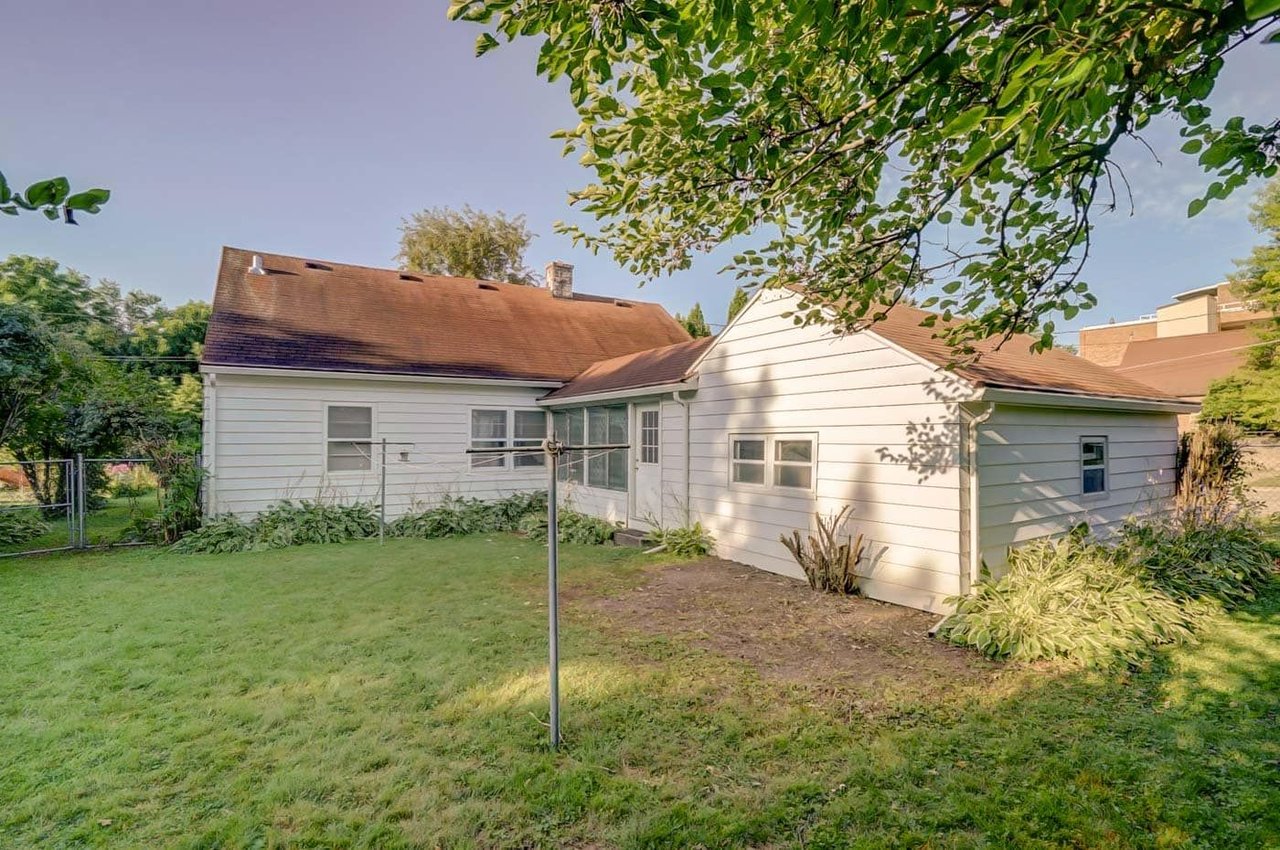 Rear of a white house with a brown roof, garden beds, and a clothesline across the lawn.