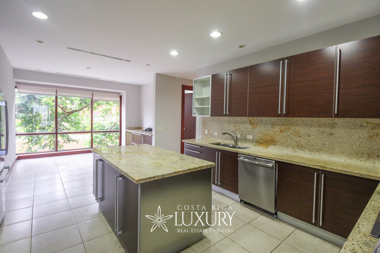 Modern apartment kitchen with a granite island, dark wood cabinets, and large windows overlooking greenery.