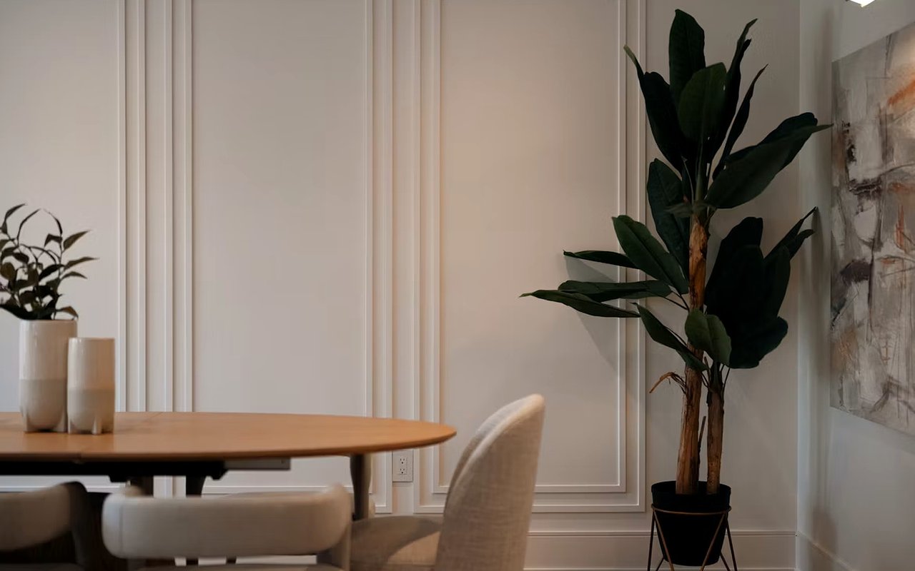 Dining area with a round wooden table, upholstered chairs, and tall potted plant.