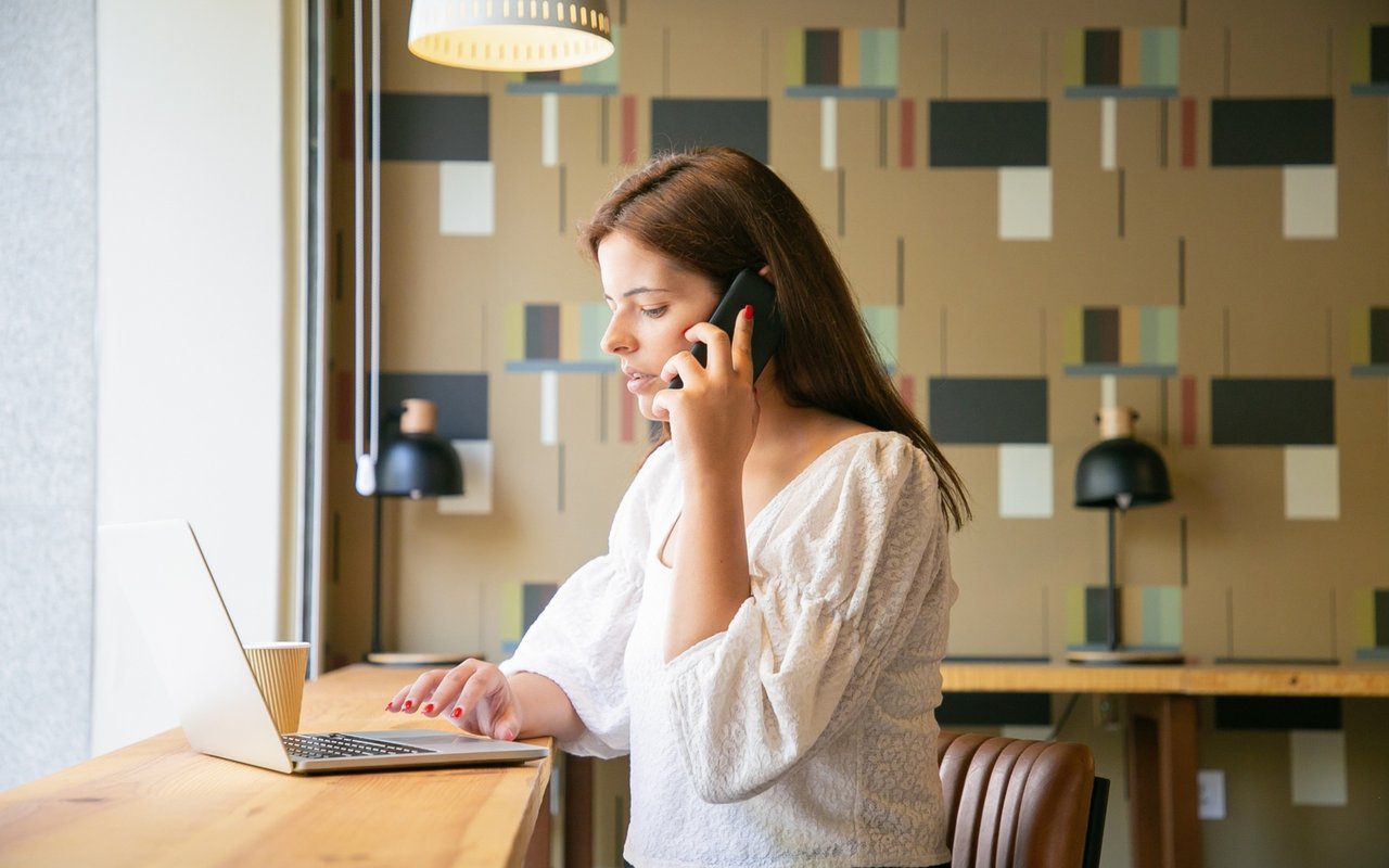 Woman talks on her phone while using a laptop at a wooden counter.