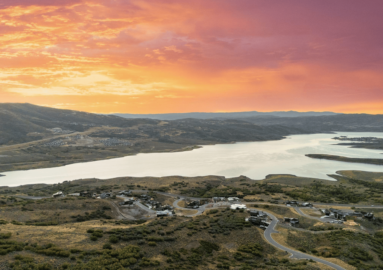 Aerial view of a lakeside community at sunset, with winding roads, homes, and hills across the water.