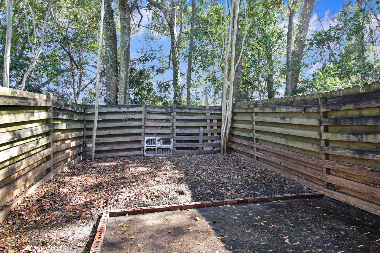 Wooden fenced corner of a backyard with trees, fallen leaves, and two wire gate panels leaning against the fence. The atmosphere is serene and natural.